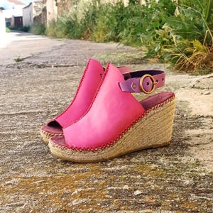 May include: Bright pink wedge sandals with a woven jute sole and a purple ankle strap. The sandals have a peep-toe design and are accented with red stitching. The buckle is gold-colored.