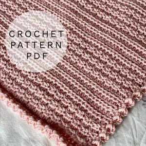 Könnte beinhalten: Nahaufnahme einer rosa gehäkelten Decke mit einem strukturierten, gerippten Design. Die Decke hat einen gewellten Rand und den Text "CROCHET PATTERN PDF" in einem weißen Kreis. Die Decke liegt auf einer Oberfläche mit floralem Muster.