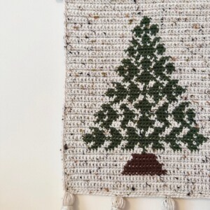May include: A handmade, crocheted wall hanging featuring a green Christmas tree design on a speckled, off-white background. The tree has a brown trunk. The wall hanging is finished with tassels and a wooden dowel for hanging.