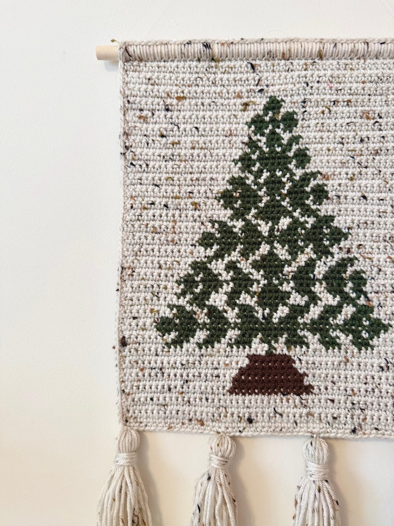 May include: A handmade, crocheted wall hanging featuring a green Christmas tree design on a speckled, off-white background. The tree has a brown trunk. The wall hanging is finished with tassels and a wooden dowel for hanging.