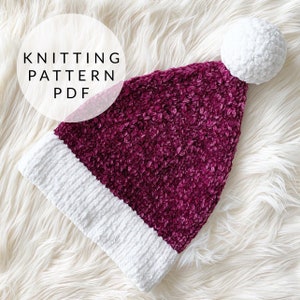 Può includere: Un cappello da Babbo Natale di lana color bordeaux e bianco con un pon pon bianco in cima. Il cappello è su una superficie bianca e soffice.  KNITTING PATTERN PDF.
