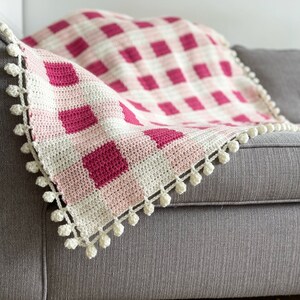Pode incluir: Uma manta de croch&ecirc; em gingham rosa e branco com uma borda de pompons brancos.