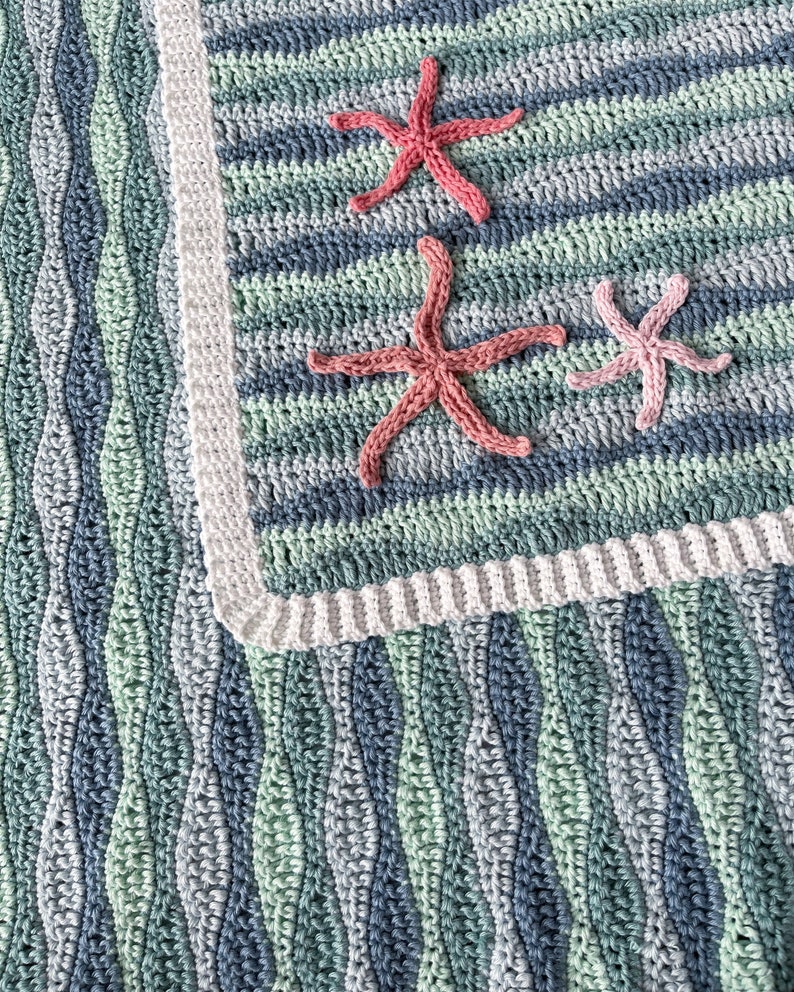 K&ouml;nnte beinhalten: Eine geh&auml;kelte Babydecke mit einem wellenf&ouml;rmigen Muster in Blaut&ouml;nen und Gr&uuml;nt&ouml;nen. Die Decke hat einen wei&szlig;en Rand und ist mit drei geh&auml;kelten Seesternen in Rosa und Rot verziert.