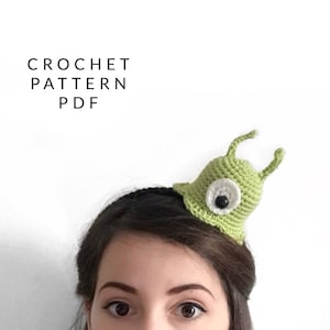 Puede incluir: Una diadema de babosa de ganchillo verde con un ojo blanco y una pupila negra. La diadema la lleva una persona con el pelo castaño.