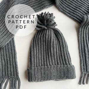May include: A gray crocheted beanie with a pom pom on top and a matching gray crocheted scarf. The scarf has fringe on the ends. The image includes the text "CROCHET PATTERN PDF".