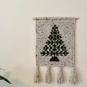 May include: A handmade crochet wall hanging featuring a dark green Christmas tree design on a speckled beige background. The tree has a brown trunk. The wall hanging is finished with four cream-colored tassels and a wooden dowel.