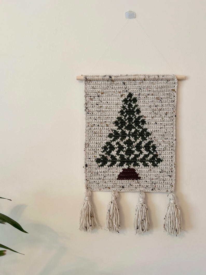 May include: A handmade crochet wall hanging featuring a dark green Christmas tree design on a speckled beige background. The tree has a brown trunk. The wall hanging is finished with four cream-colored tassels and a wooden dowel.