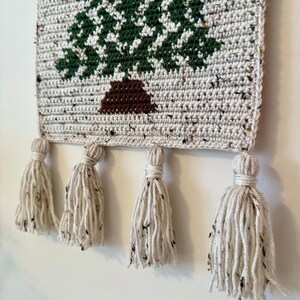 May include: Crocheted wall hanging featuring a green and white tree design with a brown trunk. The rectangular piece is made of speckled yarn and has four cream-colored tassels hanging from the bottom. The wall hanging is a decorative item.