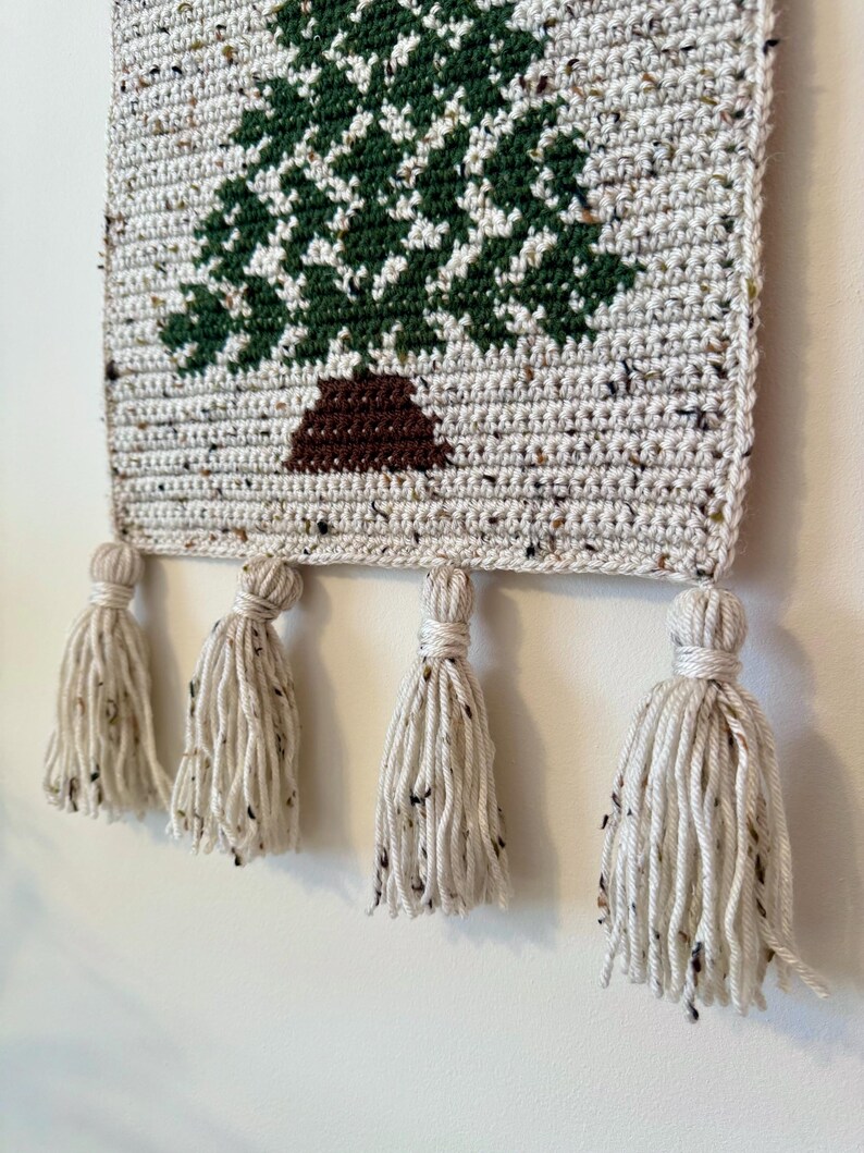 May include: Crocheted wall hanging featuring a green and white tree design with a brown trunk. The rectangular piece is made of speckled yarn and has four cream-colored tassels hanging from the bottom. The wall hanging is a decorative item.