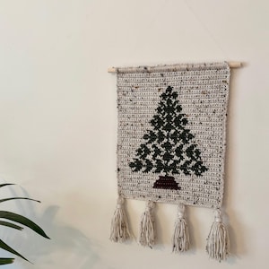 May include: Crocheted wall hanging featuring a Christmas tree design. The tree is dark green with a brown base, set against a speckled beige background. The piece is finished with four cream-colored tassels and a wooden dowel for hanging.