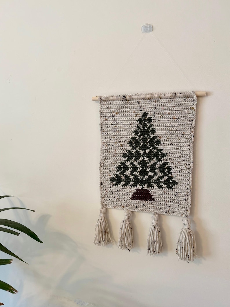 May include: Crocheted wall hanging featuring a Christmas tree design. The tree is dark green with a brown base, set against a speckled beige background. The piece is finished with four cream-colored tassels and a wooden dowel for hanging.
