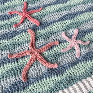 K&ouml;nnte beinhalten: Eine geh&auml;kelte Babydecke mit einem gestreiften Muster in Blau, Gr&uuml;n und Wei&szlig;. Drei rosafarbene geh&auml;kelte Seesterne sind an der Decke befestigt.