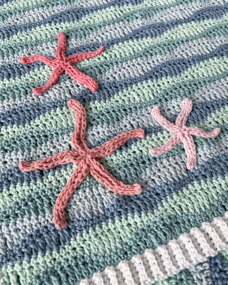 K&ouml;nnte beinhalten: Eine geh&auml;kelte Babydecke mit einem gestreiften Muster in Blau, Gr&uuml;n und Wei&szlig;. Drei rosafarbene geh&auml;kelte Seesterne sind an der Decke befestigt.
