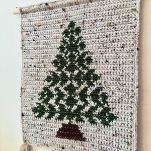 May include: A handmade, crocheted wall hanging featuring a green Christmas tree design on a speckled, off-white background. The tree has a brown trunk. The wall hanging is mounted on a wooden dowel and has three tassels at the bottom.