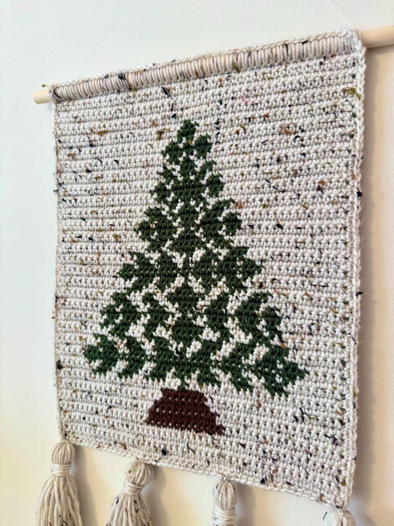 May include: A handmade, crocheted wall hanging featuring a green Christmas tree design on a speckled, off-white background. The tree has a brown trunk. The wall hanging is mounted on a wooden dowel and has three tassels at the bottom.