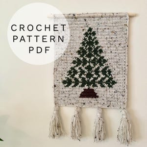 Puede incluir: Tapiz de pared tejido a ganchillo con un diseño de árbol de Navidad verde sobre un fondo gris moteado. El texto "CROCHET PATTERN PDF" está en un círculo blanco. Cuatro borlas adornan la parte inferior.