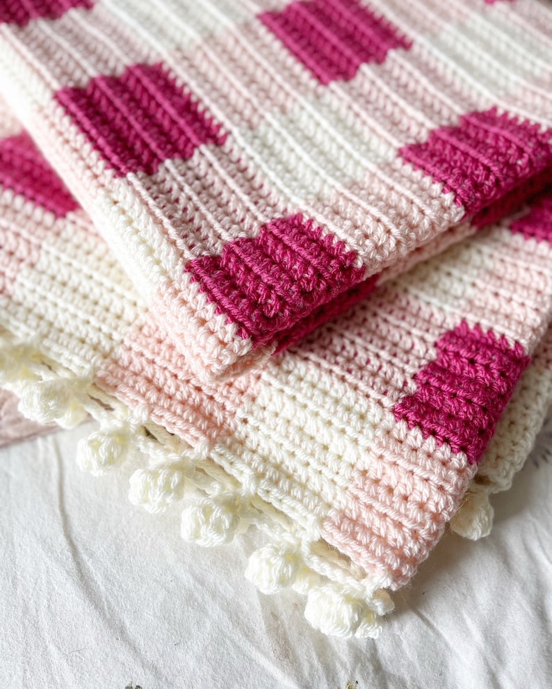Pode incluir: Um cobertor de beb&eacute; em crochet rosa e branco com uma borda em crochet branco. O cobertor tem um padr&atilde;o xadrez.