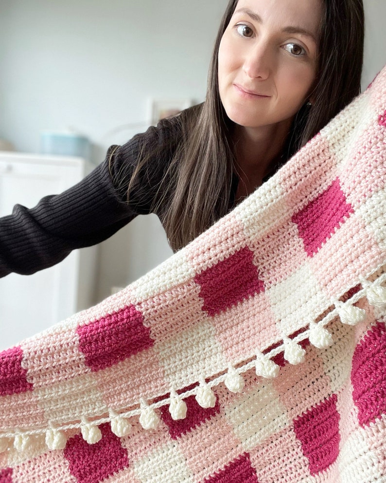 Pode incluir: Uma manta de croch&ecirc; rosa e branca com um padr&atilde;o xadrez e pompons brancos ao longo da borda.