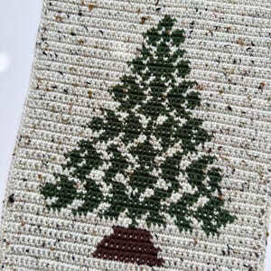 May include: A handmade, rectangular wall hanging featuring a crocheted Christmas tree design. The tree is green with a brown trunk, set against a speckled off-white background. A wooden dowel and tassels are at the top and bottom.