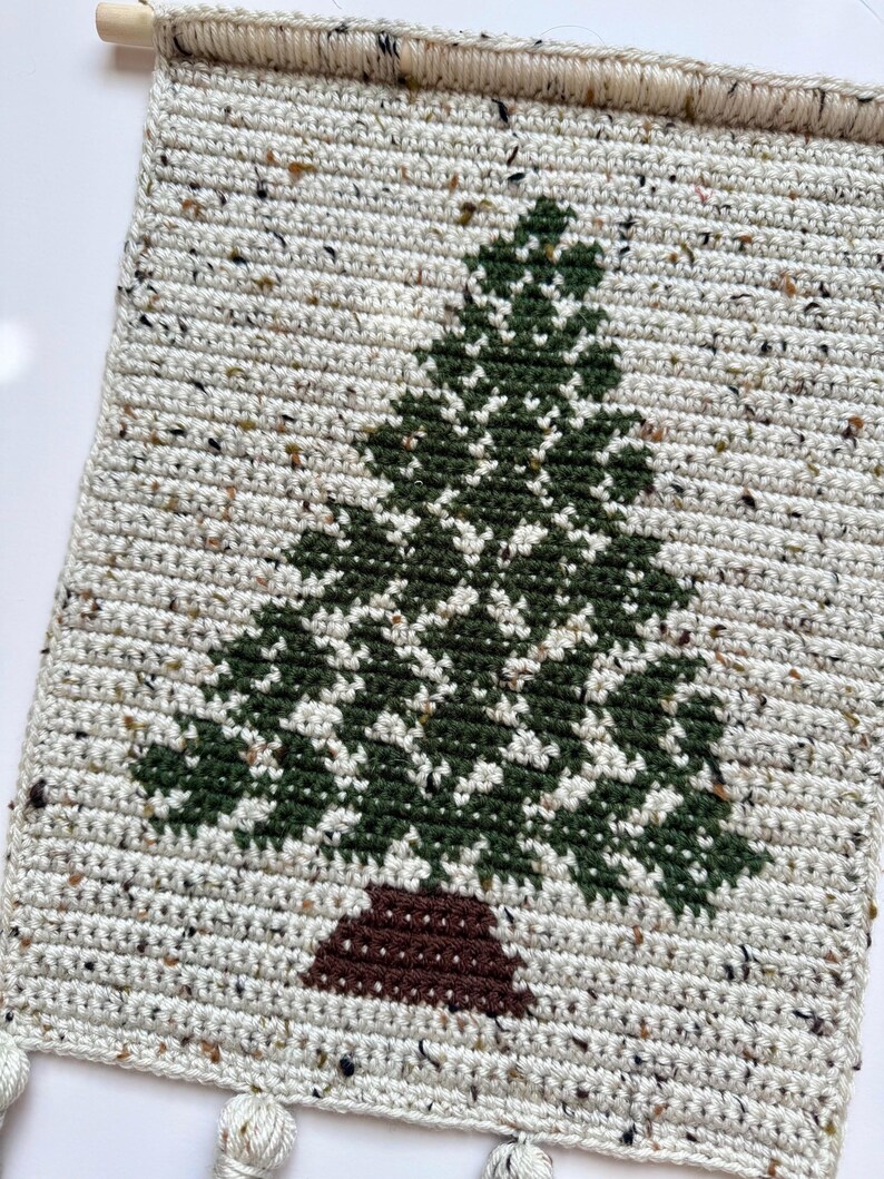 May include: A handmade, rectangular wall hanging featuring a crocheted Christmas tree design. The tree is green with a brown trunk, set against a speckled off-white background. A wooden dowel and tassels are at the top and bottom.