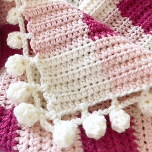 Pode incluir: Uma manta de croch&ecirc; com um padr&atilde;o listrado rosa, branco e vermelho. A manta tem uma borda de croch&ecirc; branca com pompons brancos.