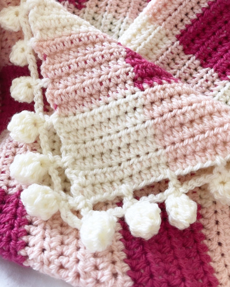 Pode incluir: Uma manta de croch&ecirc; com um padr&atilde;o listrado rosa, branco e vermelho. A manta tem uma borda de croch&ecirc; branca com pompons brancos.