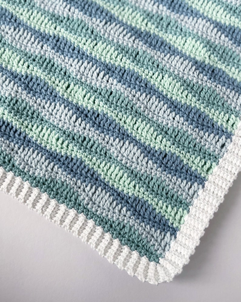 K&ouml;nnte beinhalten: Eine geh&auml;kelte Babydecke mit einem wei&szlig;en Rand und Streifen aus blauem und gr&uuml;nem Garn.