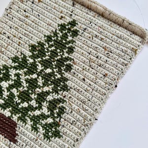 May include: Crocheted wall hanging featuring a green tree design on a speckled off-white background. The tree has a brown trunk. The piece is rectangular with a finished edge and a loop for hanging. The yarn has flecks of color.