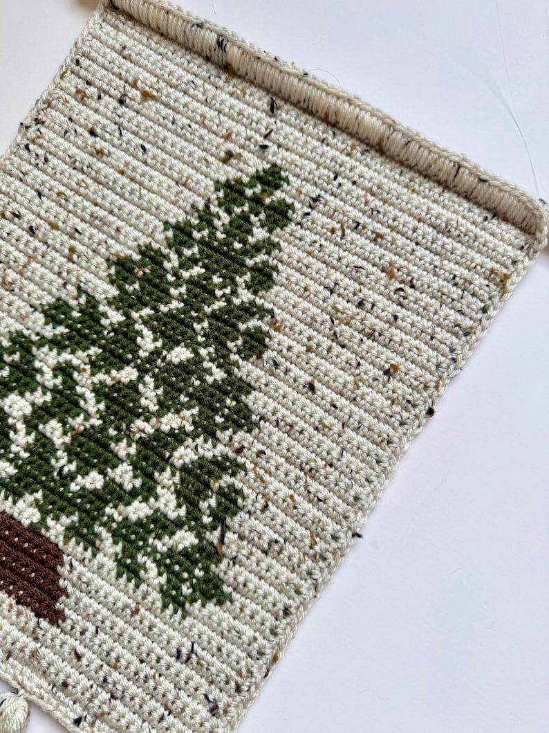 May include: Crocheted wall hanging featuring a green tree design on a speckled off-white background. The tree has a brown trunk. The piece is rectangular with a finished edge and a loop for hanging. The yarn has flecks of color.