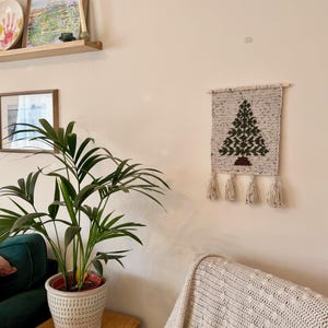 May include: A handmade, beige crochet wall hanging featuring a green Christmas tree design. The tree is detailed with dark green stitches and sits above a brown base. The wall hanging is adorned with four beige tassels.