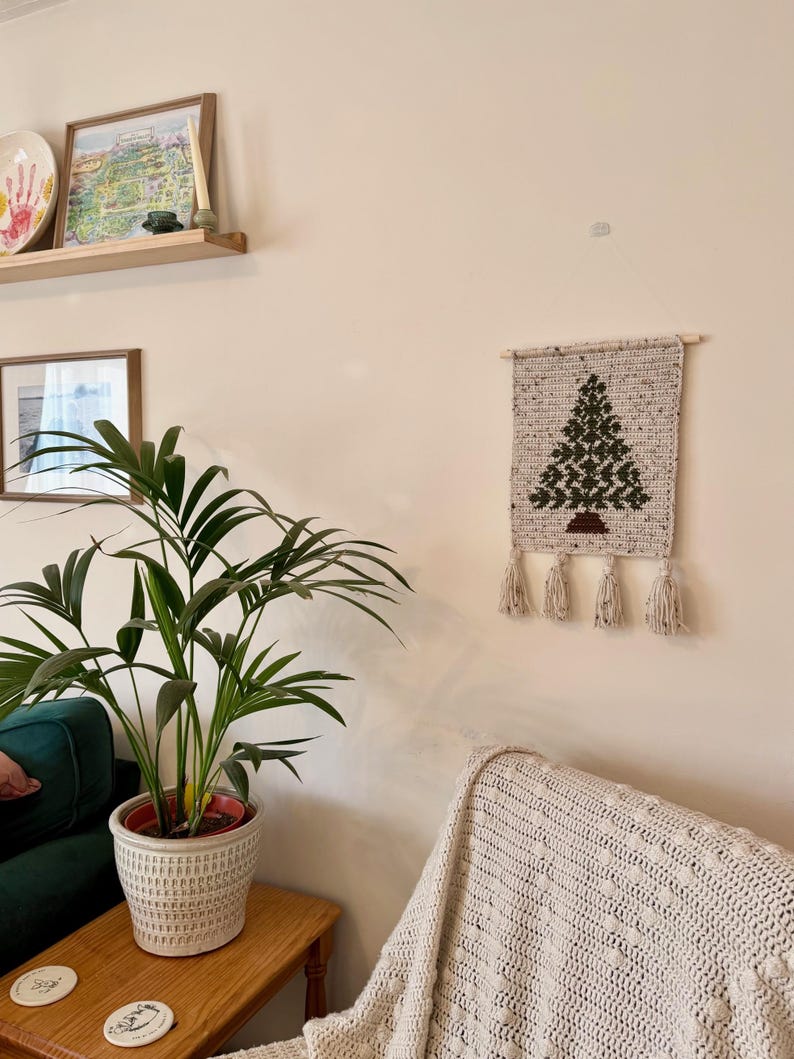May include: A handmade, beige crochet wall hanging featuring a green Christmas tree design. The tree is detailed with dark green stitches and sits above a brown base. The wall hanging is adorned with four beige tassels.