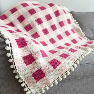 Pode incluir: Uma manta de croch&ecirc; rosa e branca com um padr&atilde;o xadrez e pompons brancos ao redor da borda. A manta est&aacute; drapeada sobre um sof&aacute; cinza.