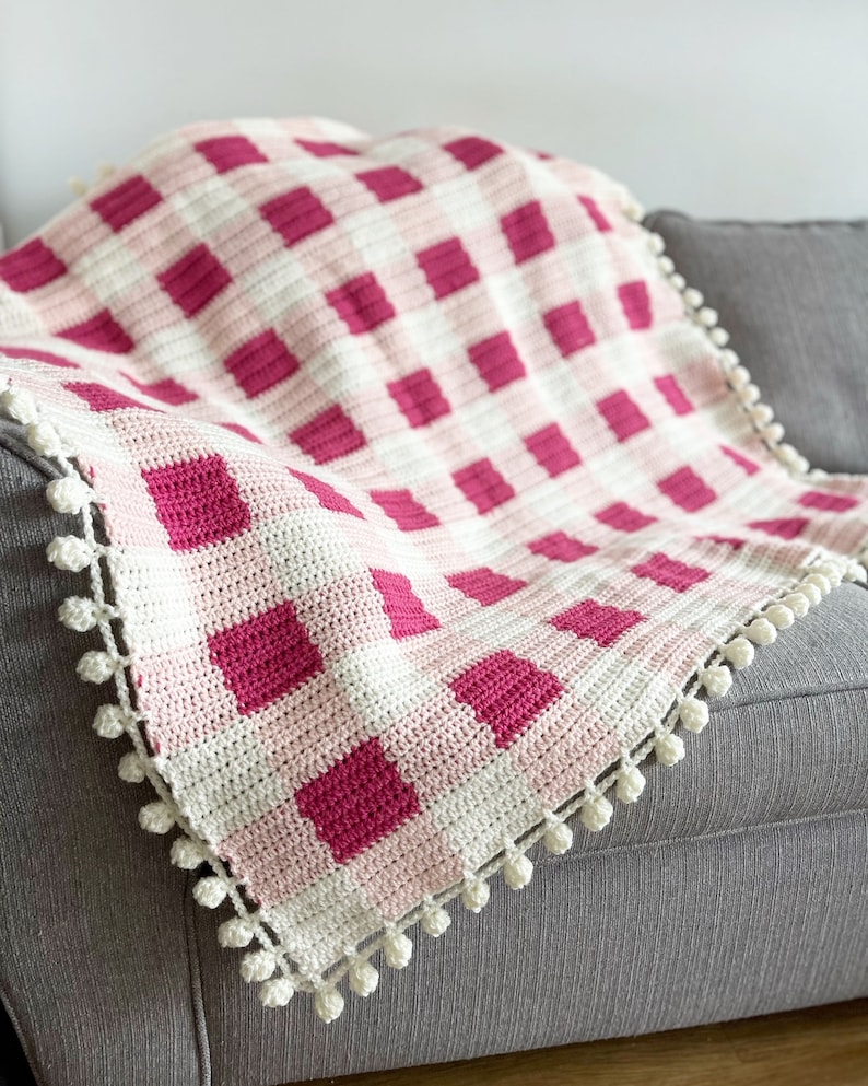 Pode incluir: Uma manta de croch&ecirc; rosa e branca com um padr&atilde;o xadrez e pompons brancos ao redor da borda. A manta est&aacute; drapeada sobre um sof&aacute; cinza.