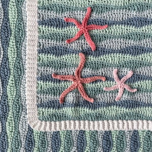 K&ouml;nnte beinhalten: Eine geh&auml;kelte Babydecke mit einem blauen, gr&uuml;nen und wei&szlig;en Wellenmuster. Die Decke hat drei rosa geh&auml;kelte Seestern-Applikationen.
