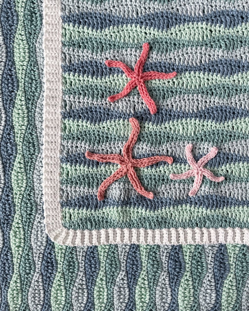 K&ouml;nnte beinhalten: Eine geh&auml;kelte Babydecke mit einem blauen, gr&uuml;nen und wei&szlig;en Wellenmuster. Die Decke hat drei rosa geh&auml;kelte Seestern-Applikationen.