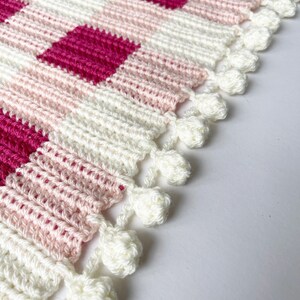 Pode incluir: Uma manta de croch&ecirc; branca com padr&atilde;o xadrez rosa e f&uacute;csia e acabamento de pompons brancos.