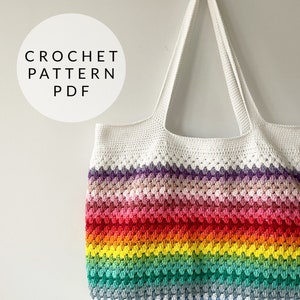 May include: A white crochet tote bag with rainbow coloured stripes. The bag has two long straps and is made with a granny stitch pattern.