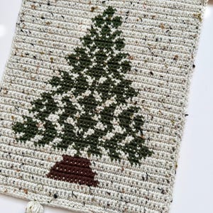 May include: A handmade, rectangular wall hanging featuring a crocheted Christmas tree design. The tree is dark green with a brown trunk, set against a speckled, off-white background. The top has a wooden dowel for hanging, and the bottom has three tassels.