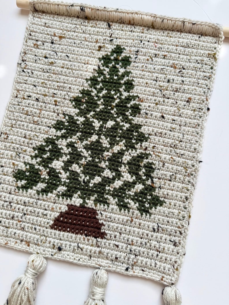 May include: A handmade, rectangular wall hanging featuring a crocheted Christmas tree design. The tree is dark green with a brown trunk, set against a speckled, off-white background. The top has a wooden dowel for hanging, and the bottom has three tassels.