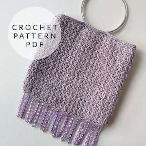 May include: A lavender crochet bag with a beaded fringe and a silver ring handle. The bag is made with a textured stitch and has a crocheted pattern. The text "CROCHET PATTERN PDF" is visible in the image.