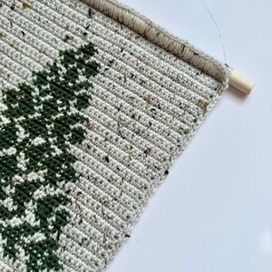 May include: Crocheted wall hanging featuring a green tree design on a speckled, off-white background. The handmade piece is mounted on a wooden dowel rod for easy display. The crochet work has a textured appearance.