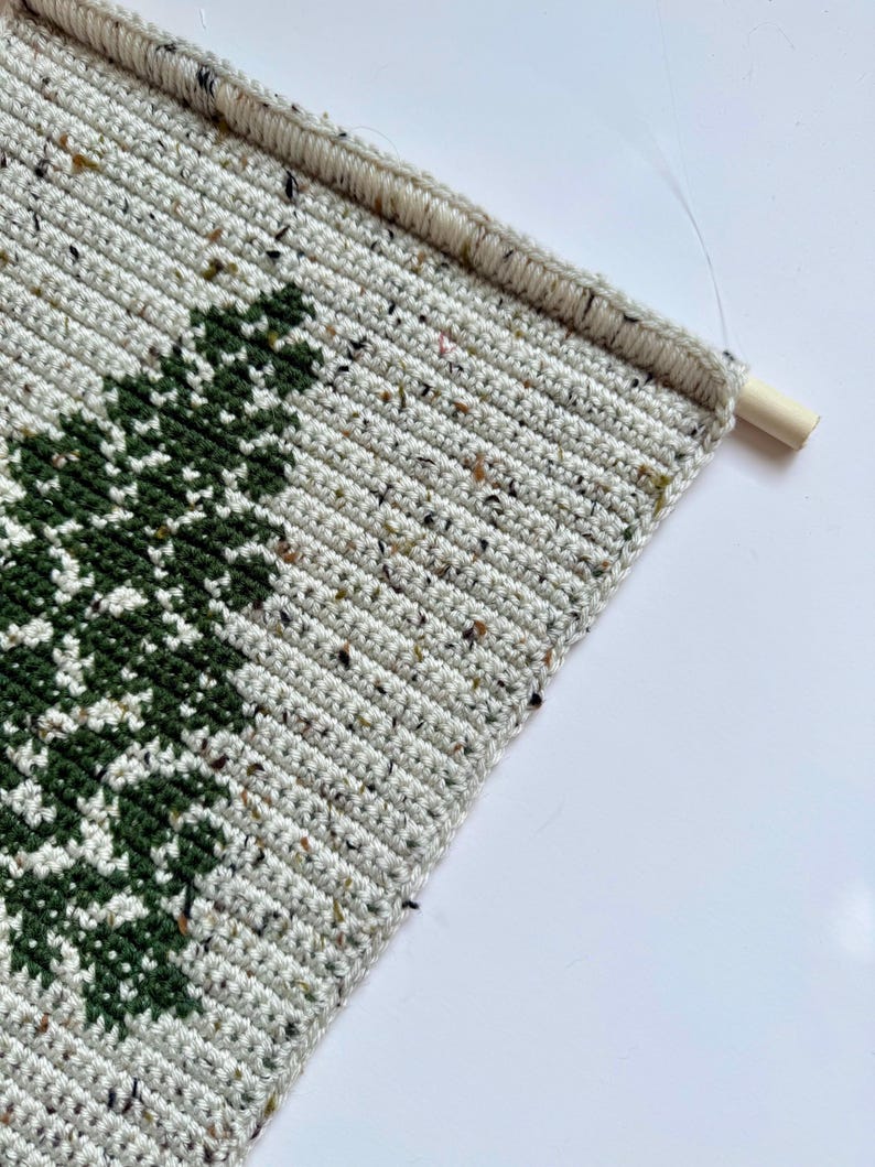 May include: Crocheted wall hanging featuring a green tree design on a speckled, off-white background. The handmade piece is mounted on a wooden dowel rod for easy display. The crochet work has a textured appearance.