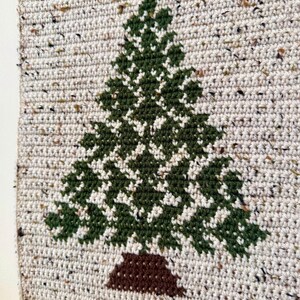 May include: A handmade, crocheted wall hanging featuring a green Christmas tree design on a speckled, off-white background. The tree has a brown trunk. The wall hanging is attached to a wooden dowel with tassels at the bottom.