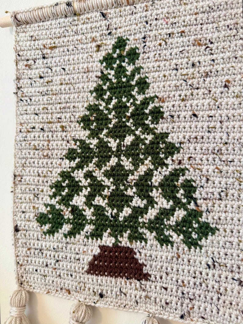 May include: A handmade, crocheted wall hanging featuring a green Christmas tree design on a speckled, off-white background. The tree has a brown trunk. The wall hanging is attached to a wooden dowel with tassels at the bottom.