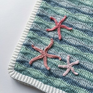 K&ouml;nnte beinhalten: Eine geh&auml;kelte Babydecke mit wei&szlig;em Rand und einem blau-gr&uuml;nen Streifenmuster. Drei rosafarbene geh&auml;kelte Seesterne sind an der Decke befestigt.