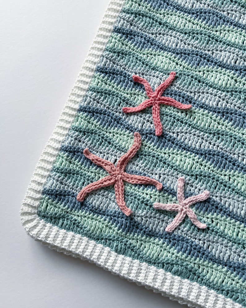 K&ouml;nnte beinhalten: Eine geh&auml;kelte Babydecke mit wei&szlig;em Rand und einem blau-gr&uuml;nen Streifenmuster. Drei rosafarbene geh&auml;kelte Seesterne sind an der Decke befestigt.