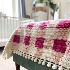 Pode incluir: Uma manta de croch&ecirc; rosa e branca com um padr&atilde;o xadrez e uma borda de pompons brancos.