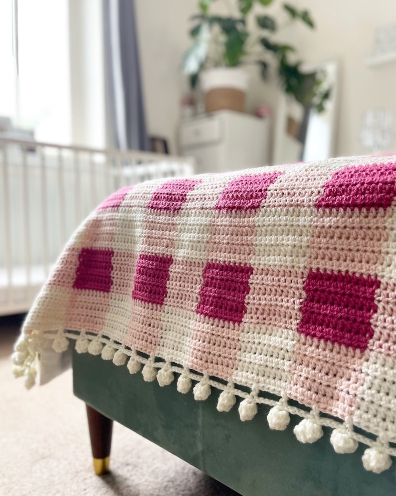 Pode incluir: Uma manta de croch&ecirc; rosa e branca com um padr&atilde;o xadrez e uma borda de pompons brancos.