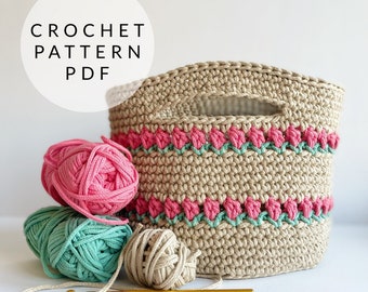May include: Crochet pattern PDF for a beige storage basket with pink and green trim. The basket is surrounded by balls of yarn in pink, green, and beige.