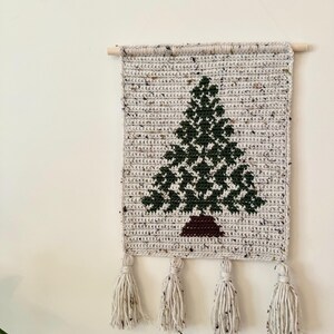 May include: A handmade, rectangular wall hanging featuring a crocheted Christmas tree design. The tree is dark green, set against a speckled gray background. The piece is finished with four cream-colored tassels and a wooden dowel for hanging.
